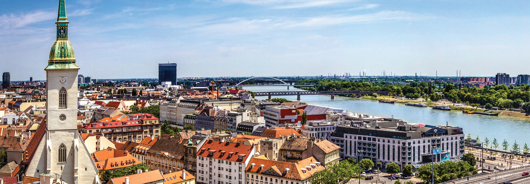 A cathedral spire overlooks the Danube River flowing through a historic Central European city with red-roofed buildings under a partly cloudy sky.