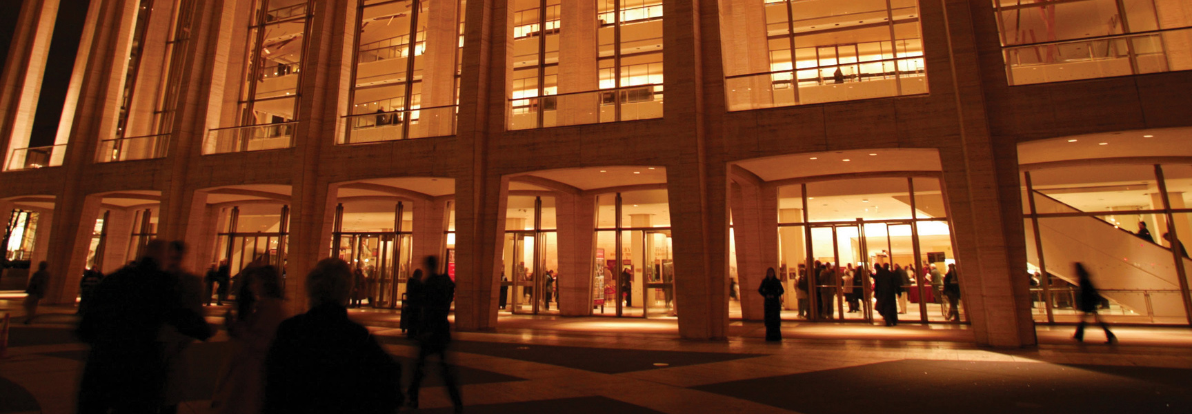 The warmly lit exterior of Lincoln Center for the Performing Arts in New York at night, with patrons walking outside the grand entrance.