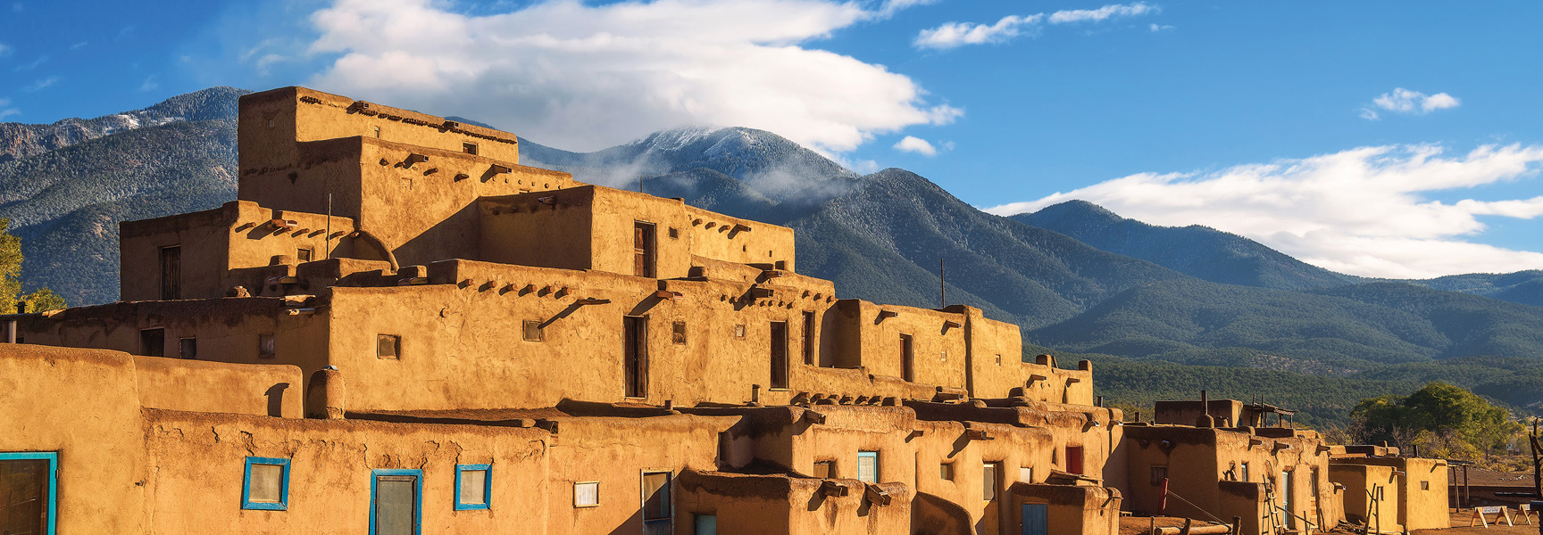 Sunlit adobe pueblo buildings in New Mexico are set against a backdrop of snow-dusted mountains under a bright blue, partly cloudy sky.
