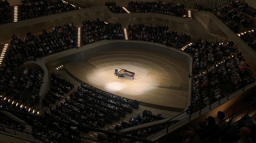 A pianist performs on a grand piano under a spotlight for a full audience inside the Elbphilharmonie concert hall in Hamburg, Germany.
