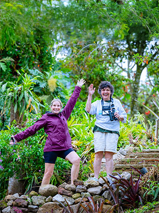 Two women smile and wave on a stone path surrounded by lush greenery and tropical plants in Costa Rica.