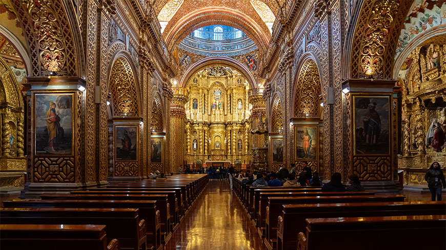 The ornate golden interior of a basilica in Quito, Ecuador, featuring intricate carvings, religious paintings, and wooden pews leading toward the altar.