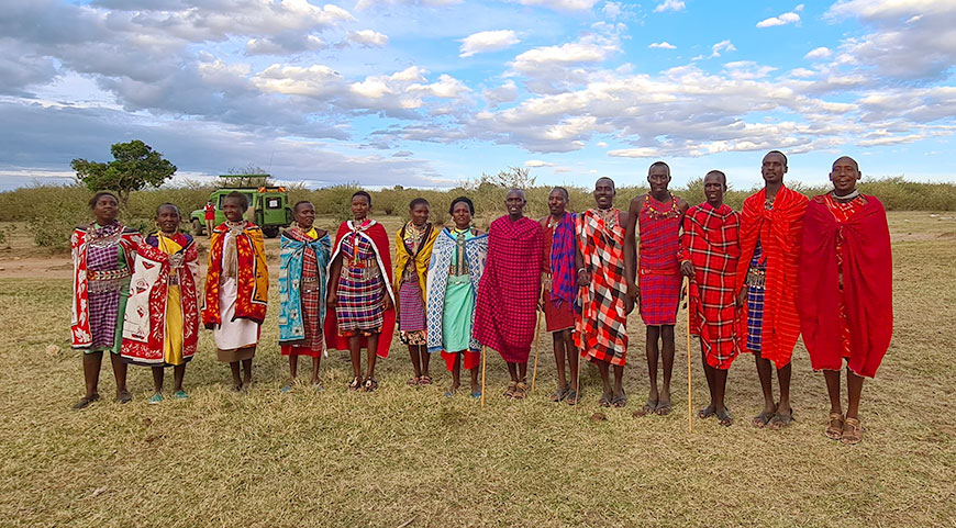 A group of Maasai men and women in colorful, traditional clothing stand together in a grassy field with a safari vehicle in the background.