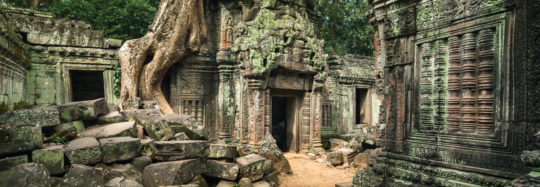 The ancient stone ruins of a temple in Southeast Asia are overgrown with large tree roots, moss, and jungle foliage.