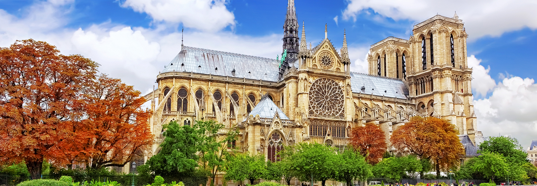 The famous Notre Dame Cathedral in Paris, France, is surrounded by bright green and orange trees under a beautiful blue sky with clouds.