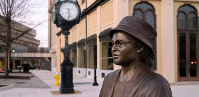 A bronze statue of civil rights activist Rosa Parks stands on a downtown sidewalk next to a vintage street clock.