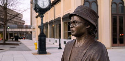 A bronze statue of civil rights activist Rosa Parks stands on a downtown sidewalk next to a vintage street clock.