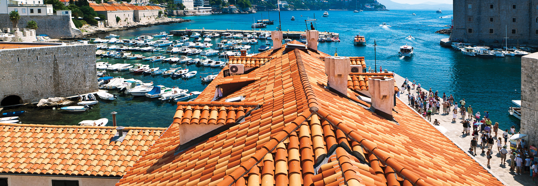 A view over the red-tiled roofs of Dubrovnik, Croatia, looking out onto the busy harbor filled with boats on a sunny day.