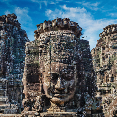 Massive stone faces carved into ancient temple towers under a blue sky, representing the history and traditions of world religions like Buddhism.