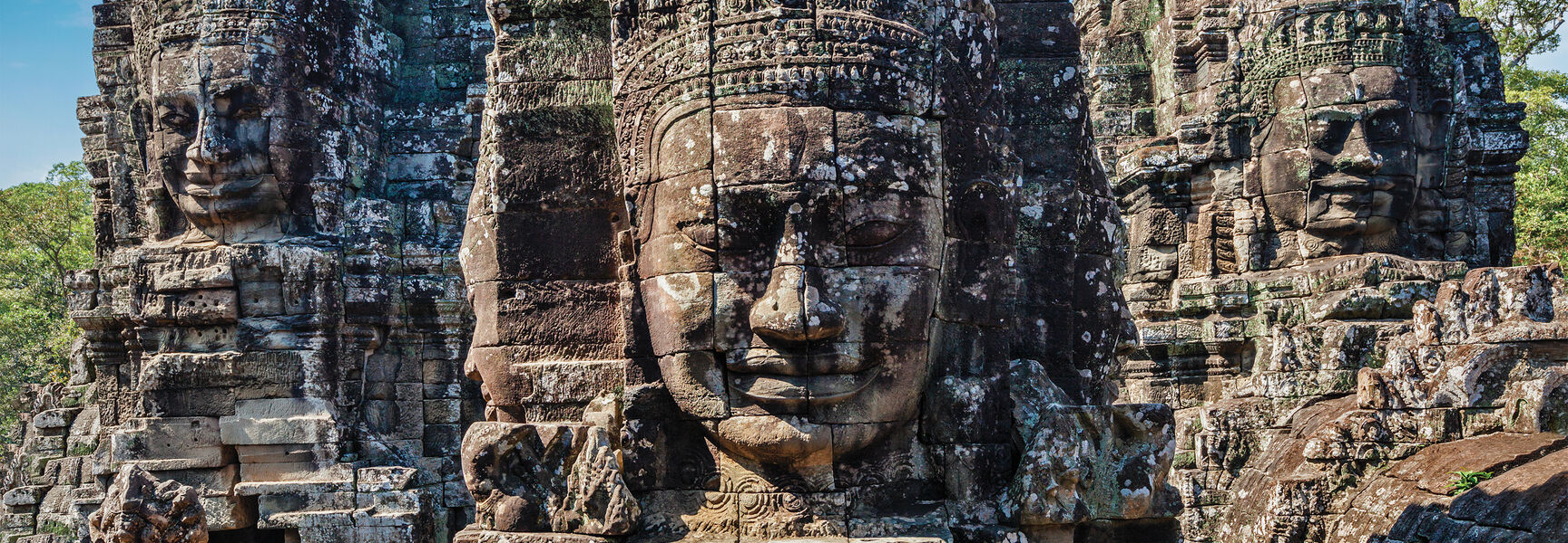 Massive stone faces carved into ancient temple towers under a blue sky, representing the history and traditions of world religions like Buddhism.