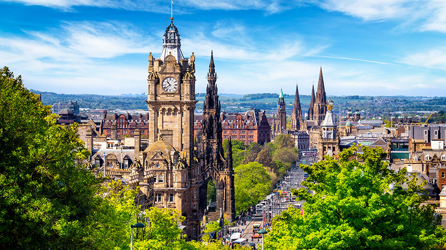 A sunny, high-angle view of the historic cityscape of Edinburgh, Scotland, with its iconic clock towers and monuments seen over bright green trees.