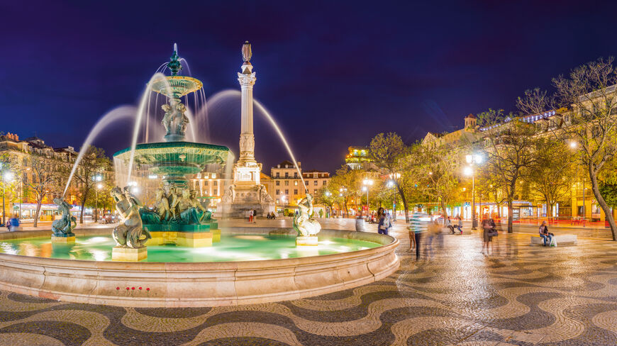 Illuminated fountain and monument in Rossio Square, Lisbon, Portugal, at night, featuring traditional wavy stone pavement.