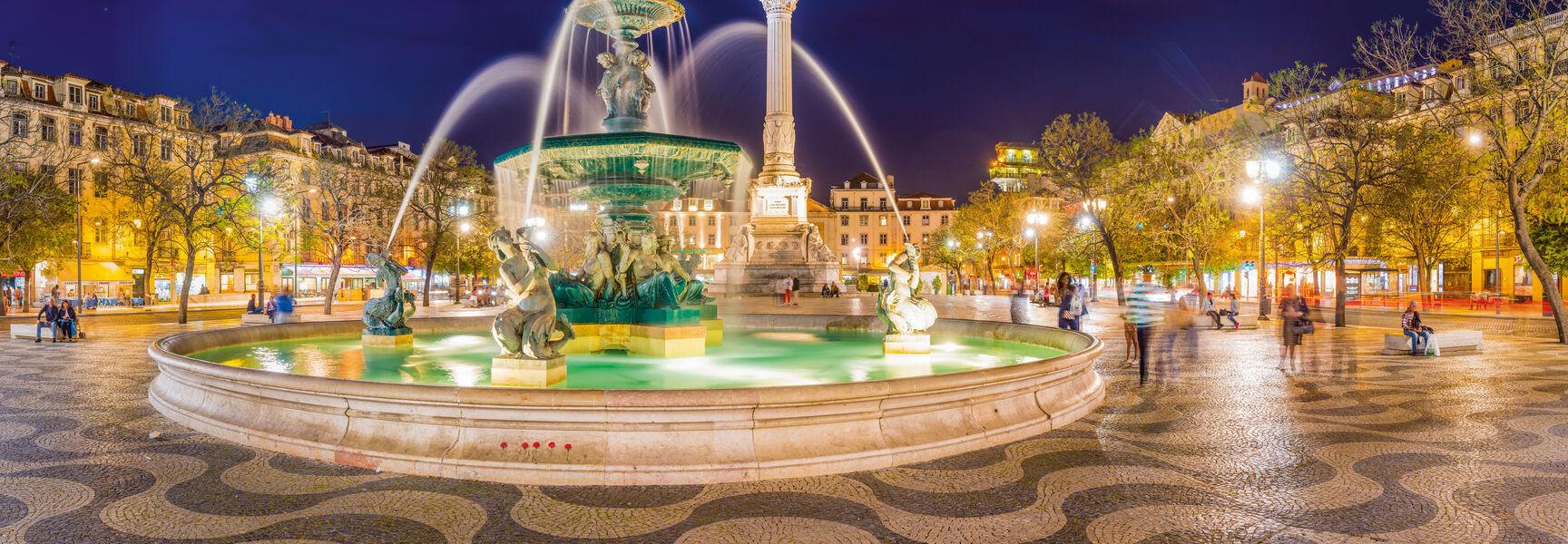 Illuminated fountain and monument in Rossio Square, Lisbon, Portugal, at night, featuring traditional wavy stone pavement.