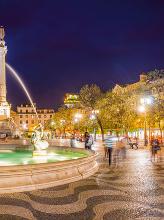 Illuminated fountain and monument in Rossio Square, Lisbon, Portugal, at night, featuring traditional wavy stone pavement.