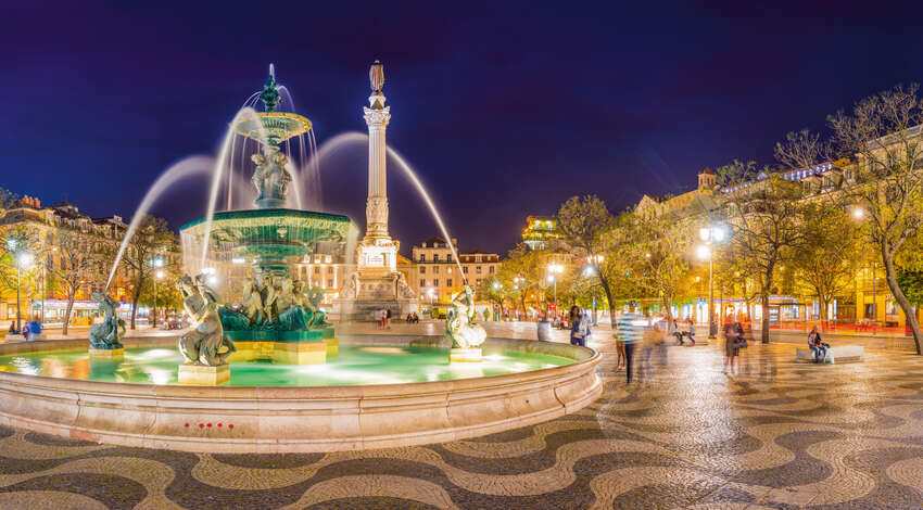 Illuminated fountain and monument in Rossio Square, Lisbon, Portugal, at night, featuring traditional wavy stone pavement.