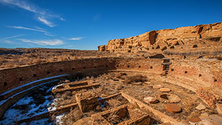The ancient stone ruins of a large, circular kiva sit in Chaco Canyon under a clear blue sky.