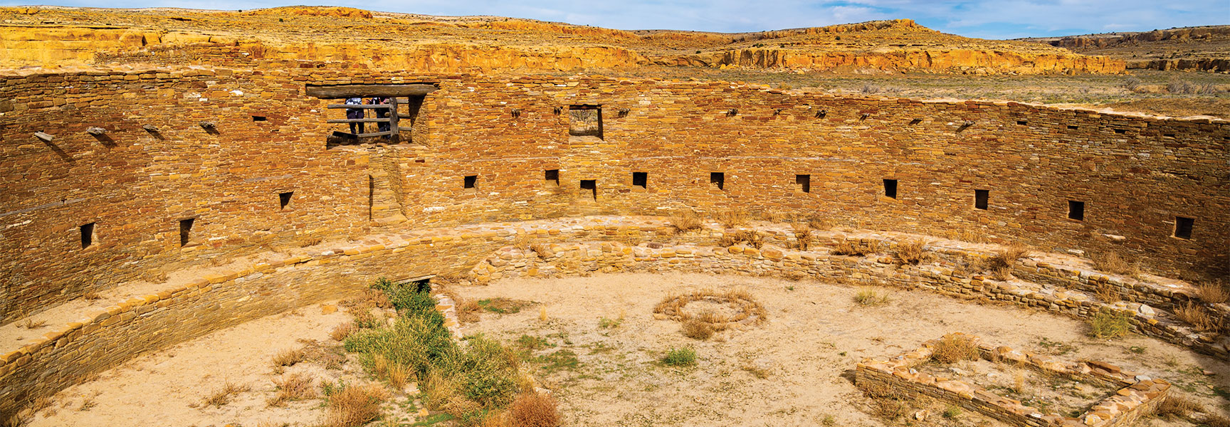 Circular stone ruins of an ancient ancestral Pueblo kiva at Chaco Canyon, New Mexico, set against a desert landscape with golden sandstone cliffs.