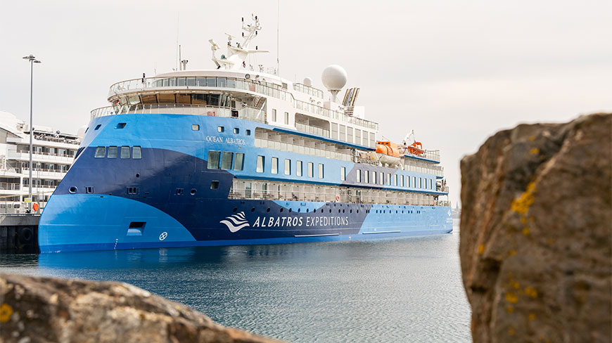 The blue and white Ocean Albatros expedition ship is docked at Scrabster Port in Scotland on a cloudy day.