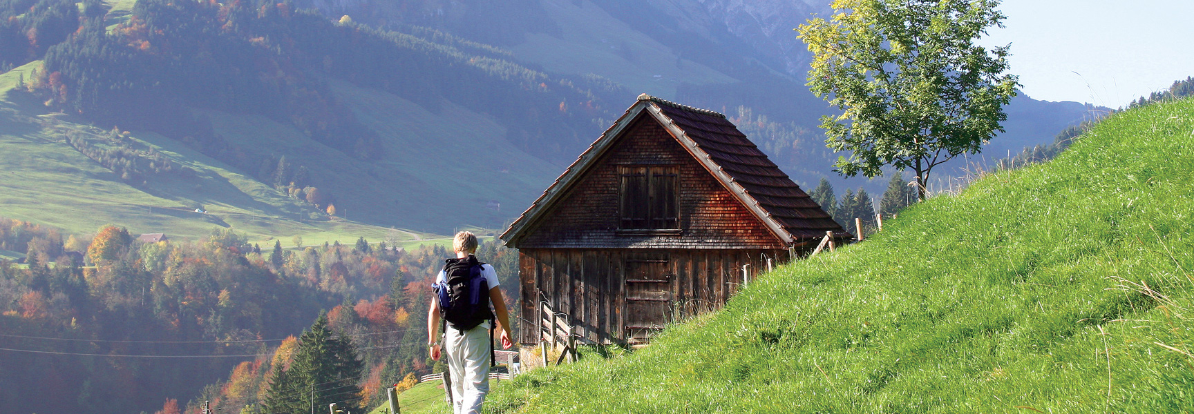 A hiker walks past a rustic wooden cabin on a grassy mountainside trail in the scenic hills of Switzerland.