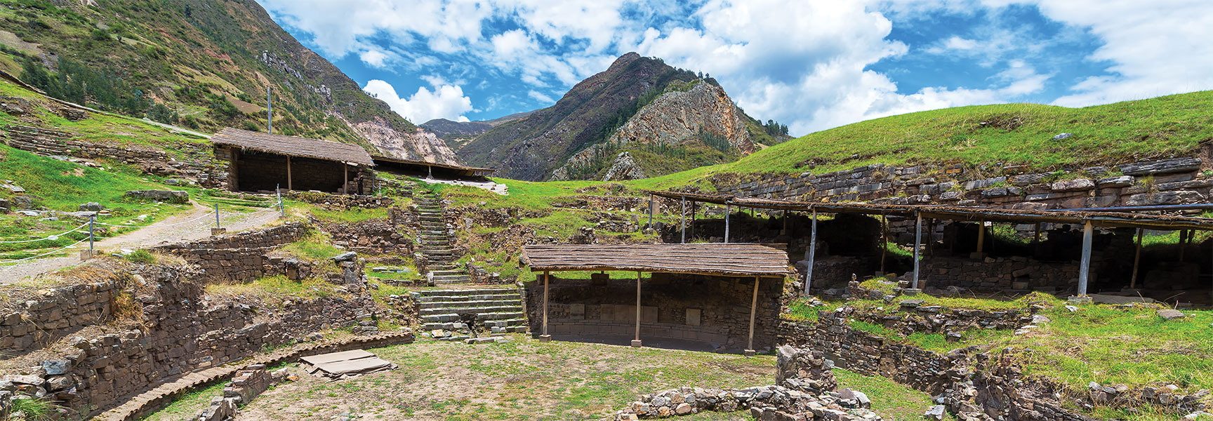 Ancient stone ruins with simple wooden roofs sit on a grassy terraced hillside at the base of a large mountain range.