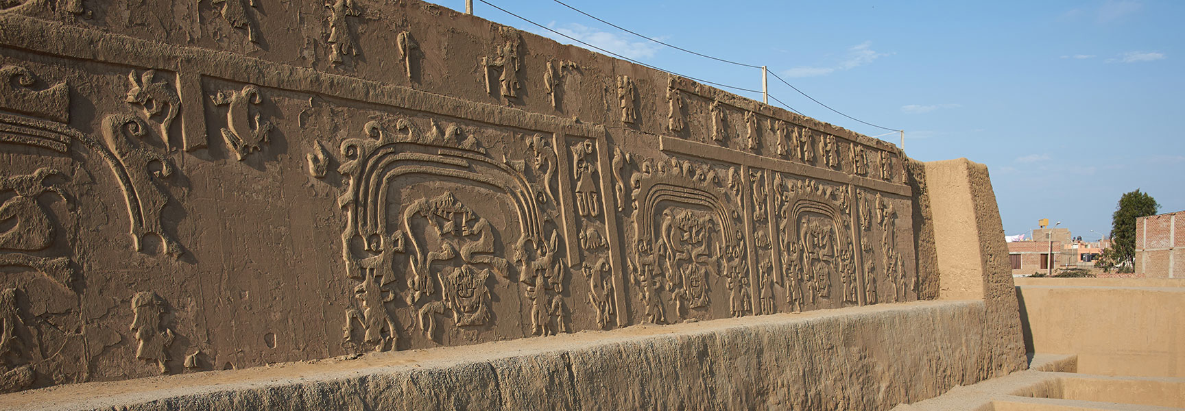 An ancient adobe wall at the Huaca del Dragón archaeological site features intricate repeating friezes under a bright blue sky.