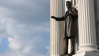A bronze statue of Abraham Lincoln stands between two large white columns with a cloudy sky in the background.