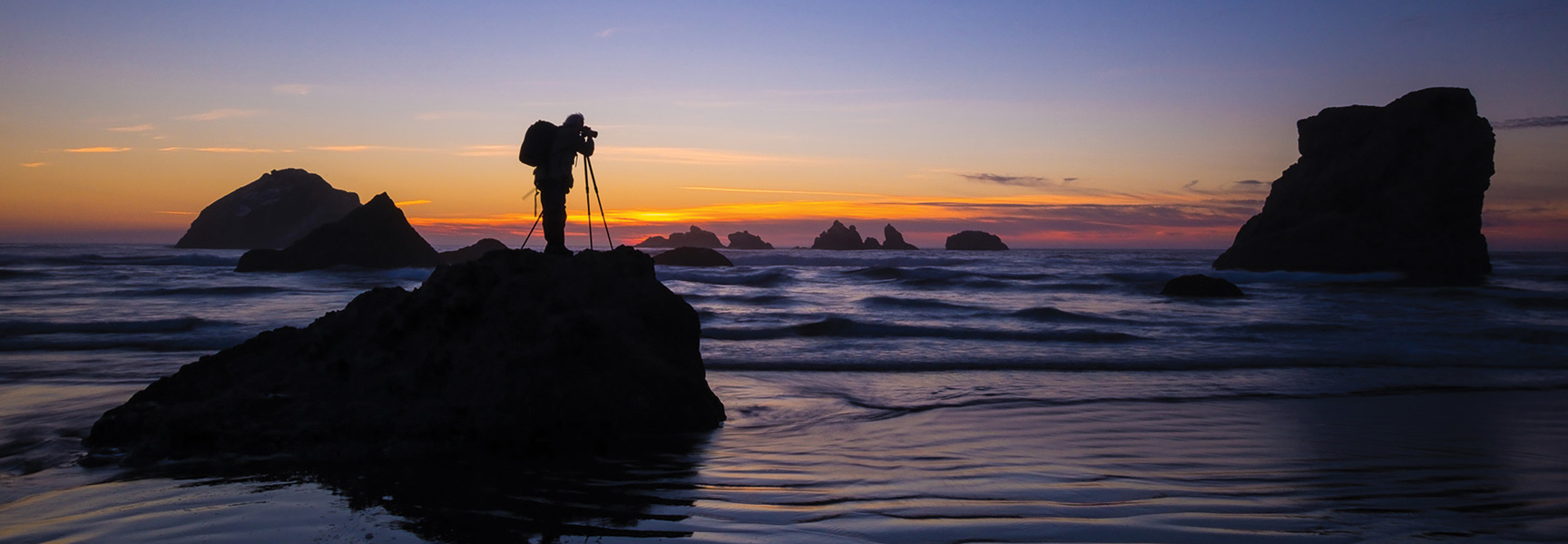 A photographer with a tripod is silhouetted against a colorful sunset on a rocky beach with sea stacks in the ocean.