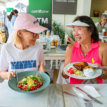 Two women smile at each other while holding plates of traditional food at a restaurant in Fira, Greece.