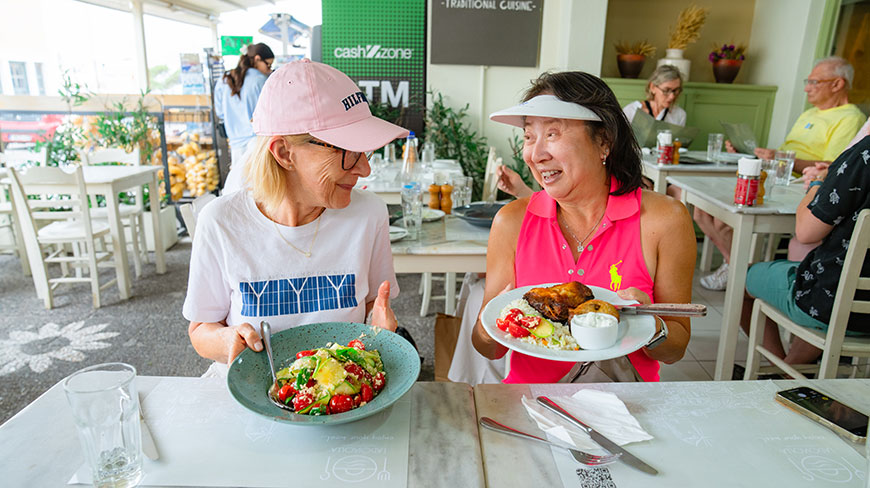 Two women smile at each other while holding plates of traditional food at a restaurant in Fira, Greece.