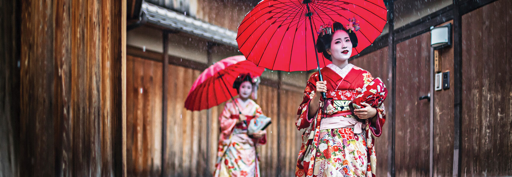 Two women in traditional red kimonos carry red umbrellas while walking through a narrow, rain-slicked street in Japan.