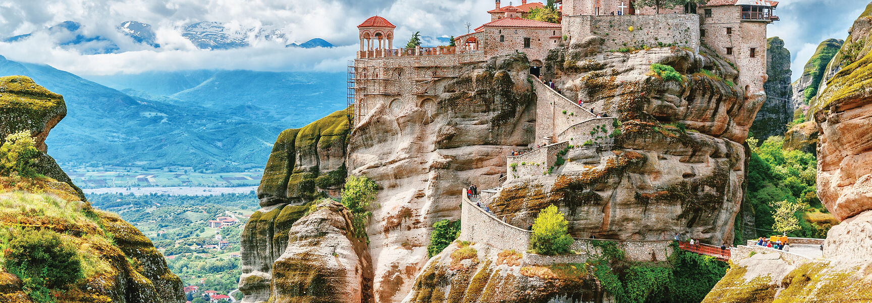 A historic Greek Orthodox monastery perched on steep rock formations in Northern Greece, with a valley and snow-capped mountains in the distance.