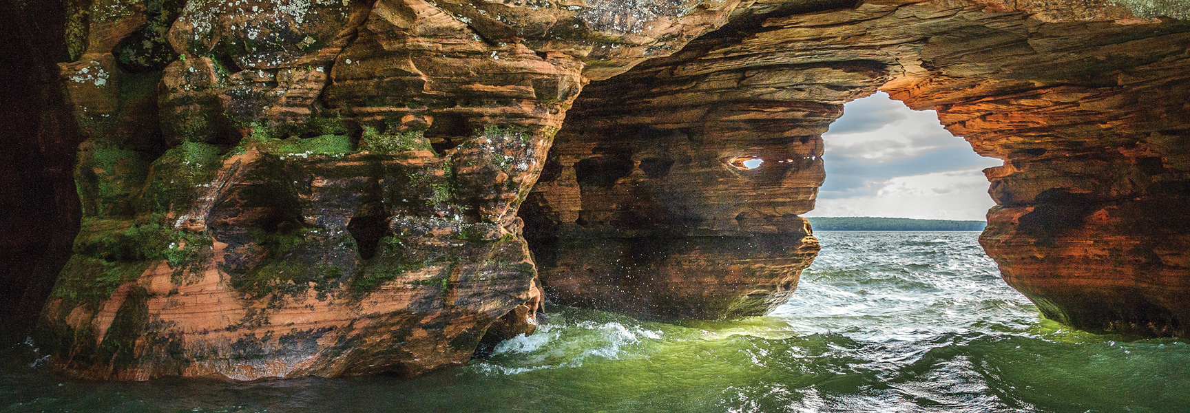 Moss-covered red rock sea caves on Lake Superior in Wisconsin with green water splashing against the formations.