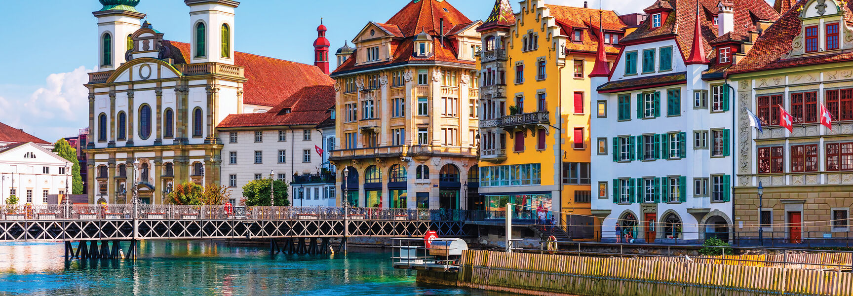 Colorful historic buildings and a twin-domed church reflect in the blue water of a stunning landscape in Switzerland.