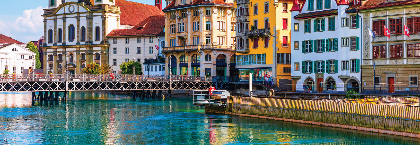 A bridge crosses a turquoise river flowing past colorful, historic buildings in a city in Switzerland.