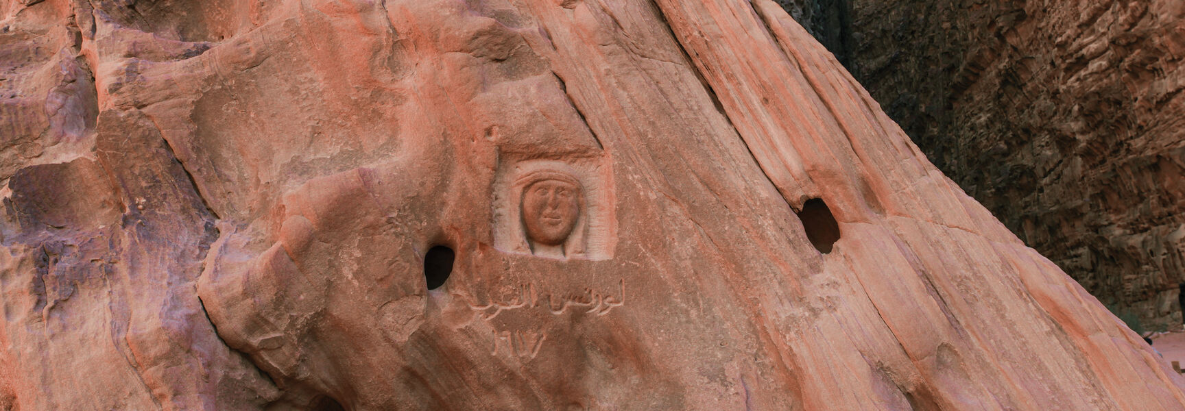 Carving of the face of Lawrence of Arabia and Arabic text on a red sandstone rock face in a desert canyon.