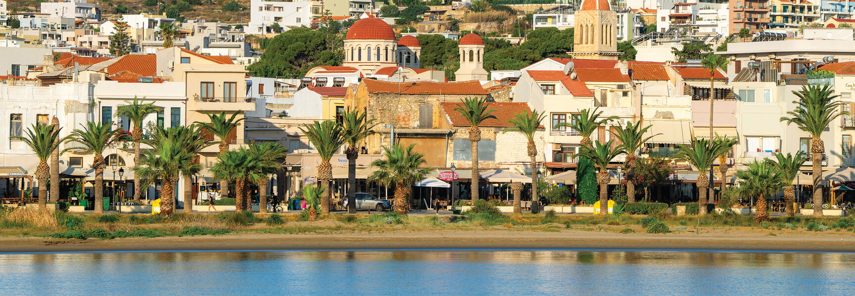 Coastal view of a Greek town with palm trees and red-roofed buildings along the blue Aegean Sea.