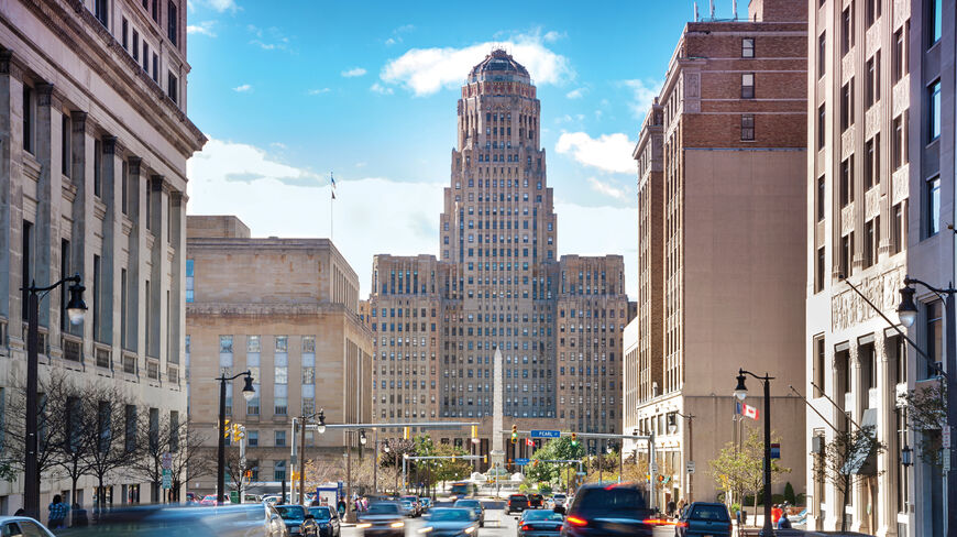 A view down a street in Buffalo, New York, toward the historic Art Deco City Hall building and McKinley Monument under a clear sky.