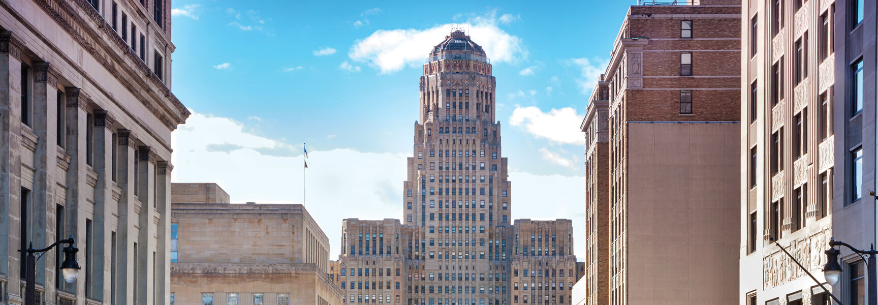 A view down a street in Buffalo, New York, toward the historic Art Deco City Hall building and McKinley Monument under a clear sky.