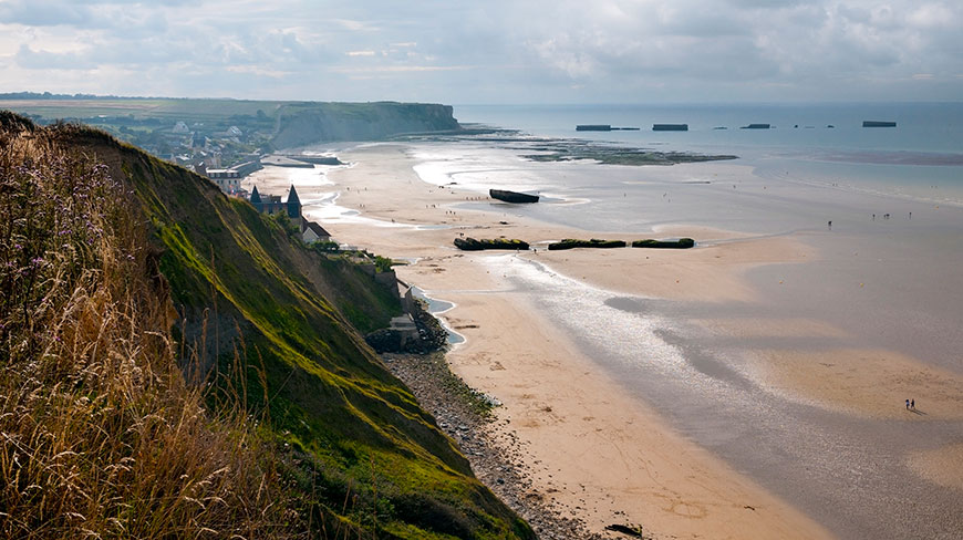 A view from the grassy cliffs over the Normandy coast shows the sandy beach and remnants of a Mulberry Harbour at low tide.