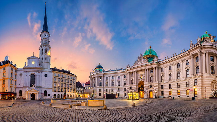The historic St. Michael's Square in Vienna, Austria, with its grand church and palace buildings illuminated against a colorful twilight sky.
