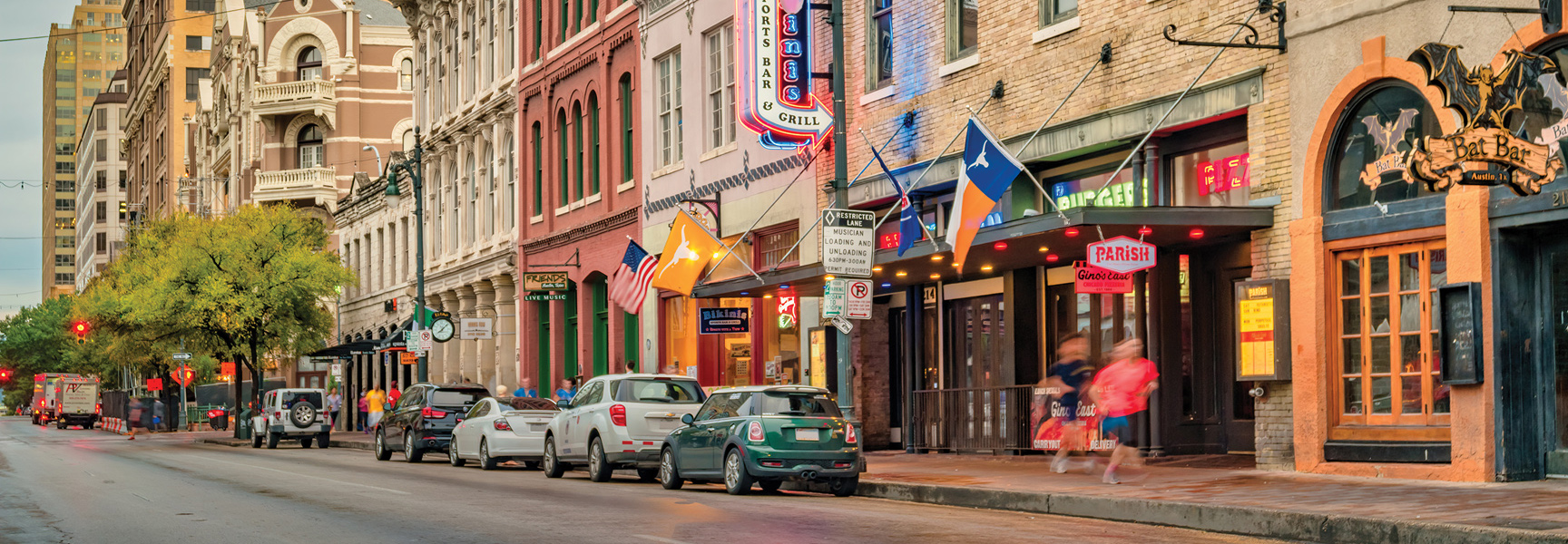 A busy street in downtown Austin, Texas, with historic buildings housing bars and restaurants, cars parked along the curb, and pedestrians on the sidewalk.