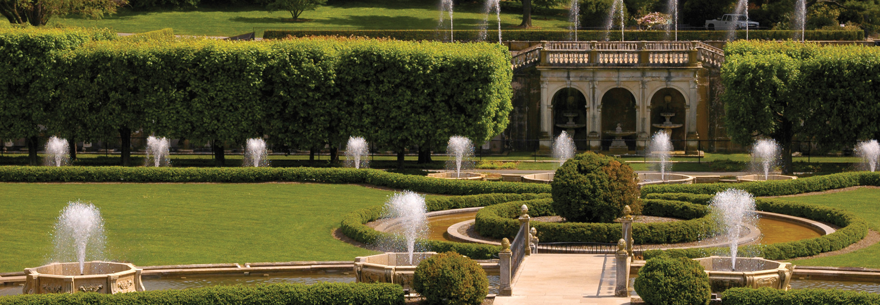 Ornate fountains spray water in a lush, formal garden with sculpted green hedges and a stone structure in Pennsylvania.