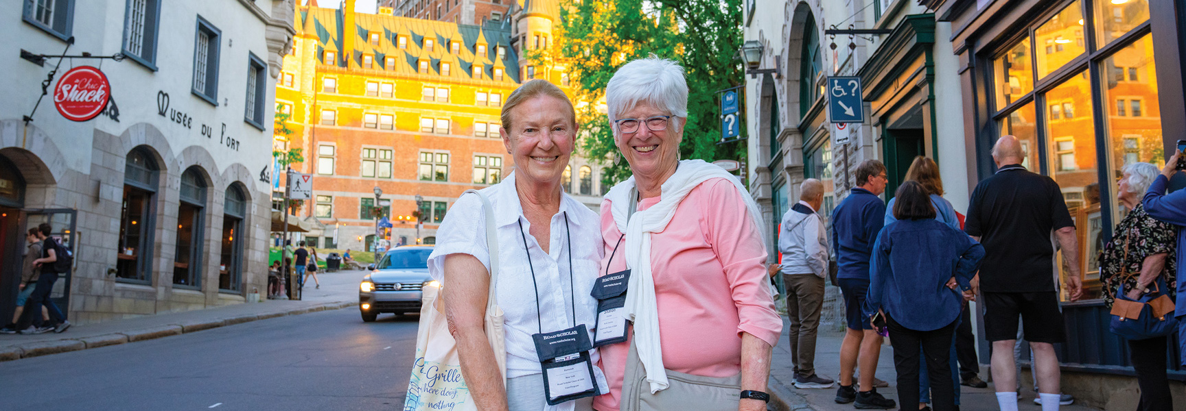 Two smiling women stand on a city street in Québec, Canada, with historic buildings and other people in the background during golden hour.