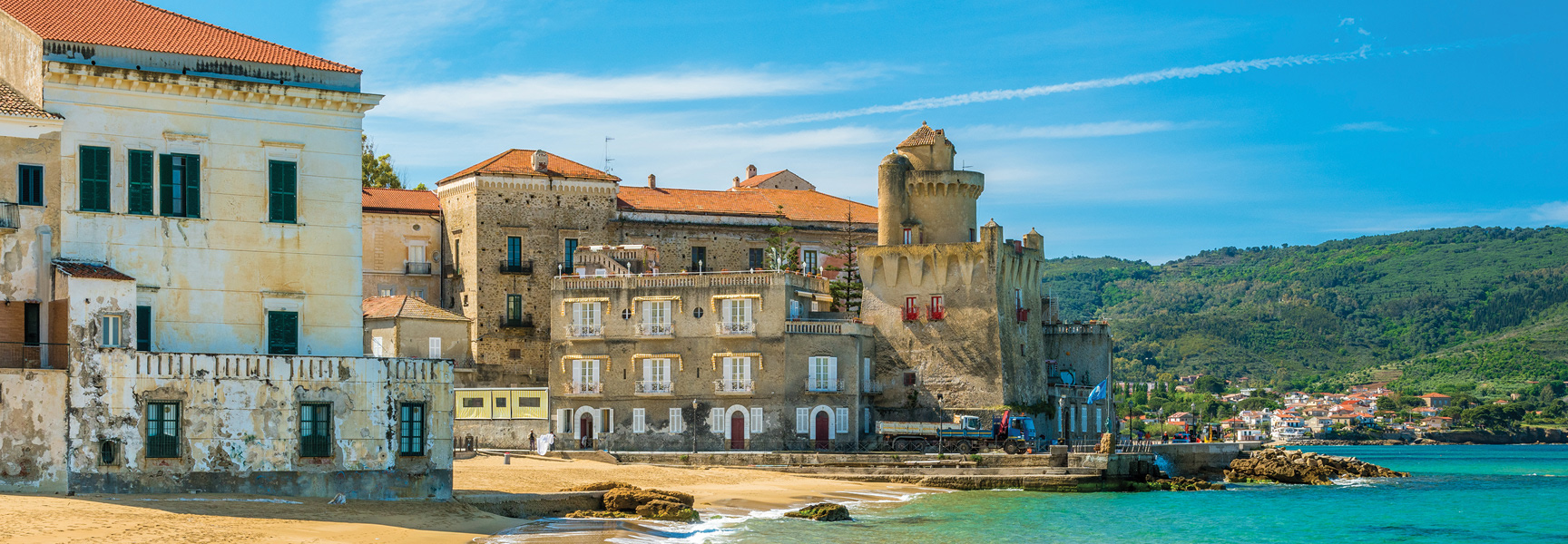A seaside castle and weathered buildings line a sandy beach along the turquoise coast of Cilento, Italy, on a sunny day.
