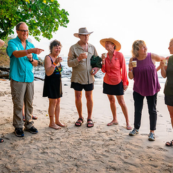 A group of travelers laugh together while enjoying drinks on a sandy beach in Costa Rica at sunset.