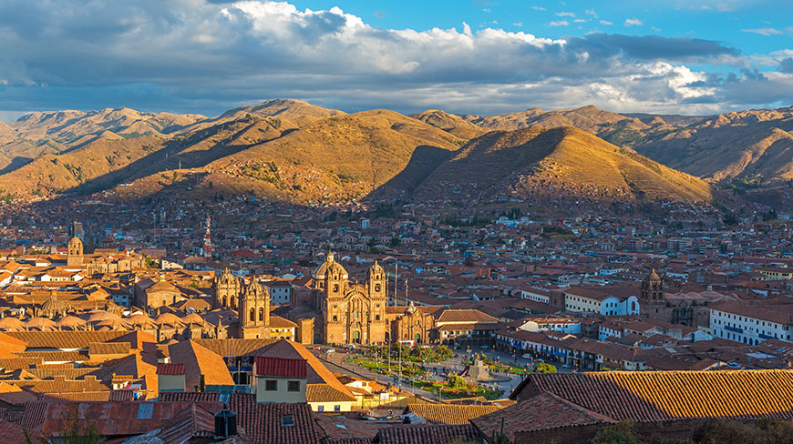 The city of Cusco, Peru, nestled in a mountain valley with its terracotta rooftops glowing in the afternoon sun under a cloudy blue sky.