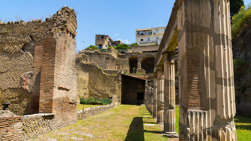 The ancient ruins of Herculaneum in Naples, Italy, show exposed brick walls and a colonnade under a bright blue sky with modern buildings above.