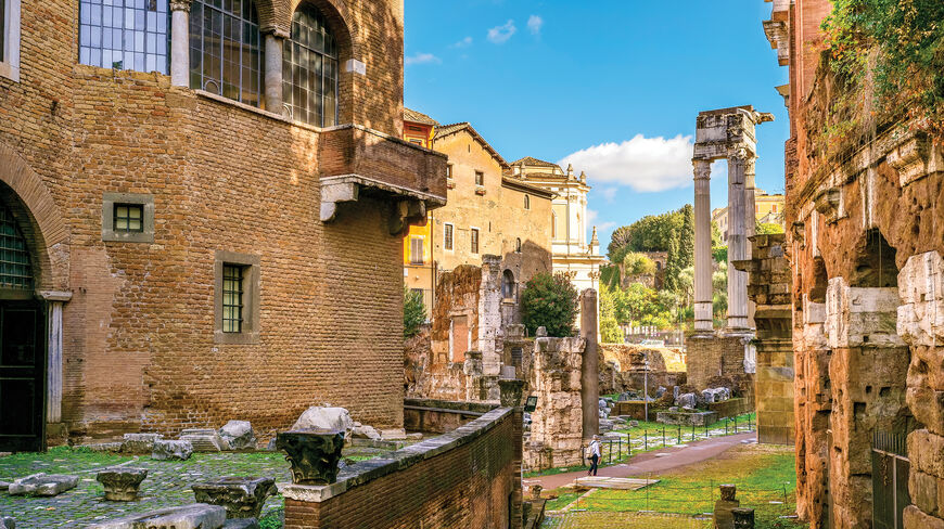 Ancient brick buildings and white marble columns of historical Roman ruins stand under a clear blue sky.