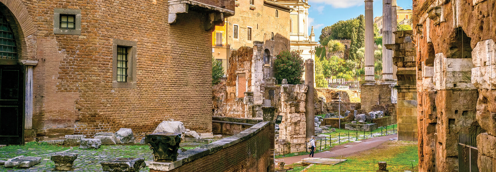Ancient brick buildings and white marble columns of historical Roman ruins stand under a clear blue sky.