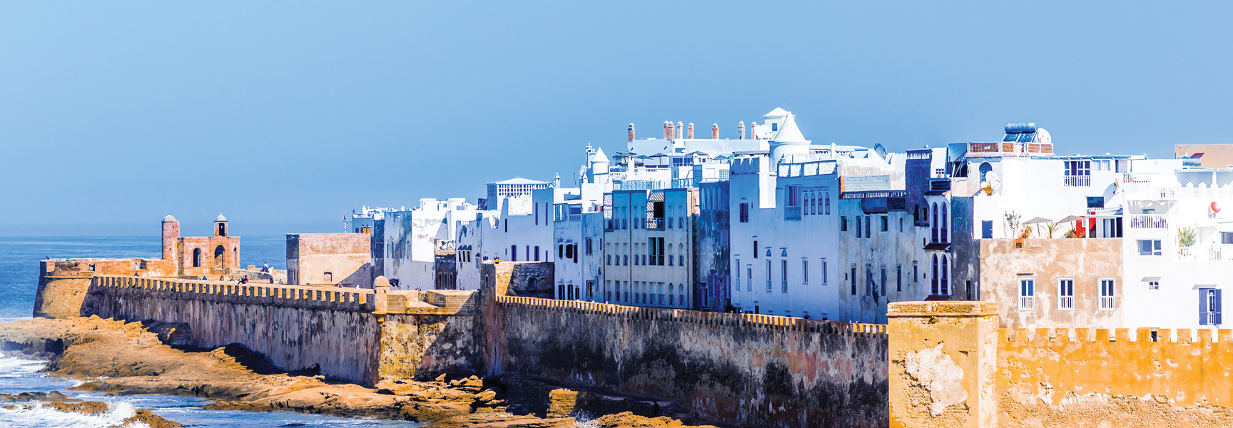 A long, stone seawall runs along the coast of Morocco, with a dense collection of white and blue buildings behind it.